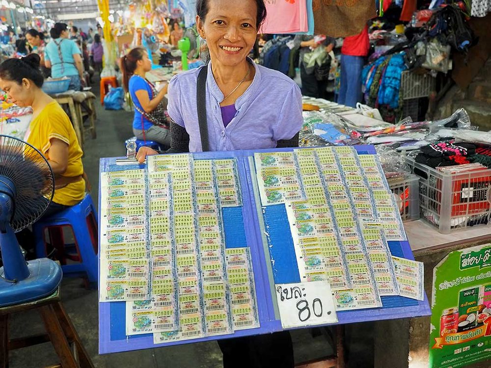 A licensed Thai Government Lottery vendor holding a board of paper lottery tickets sold in matching pairs at a Thai market